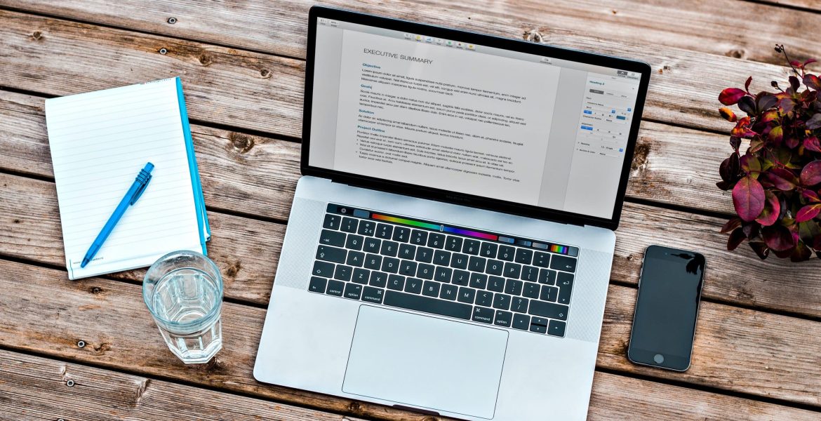 Laptop with open document beside a notebook, phone, and glass of water on a wooden table